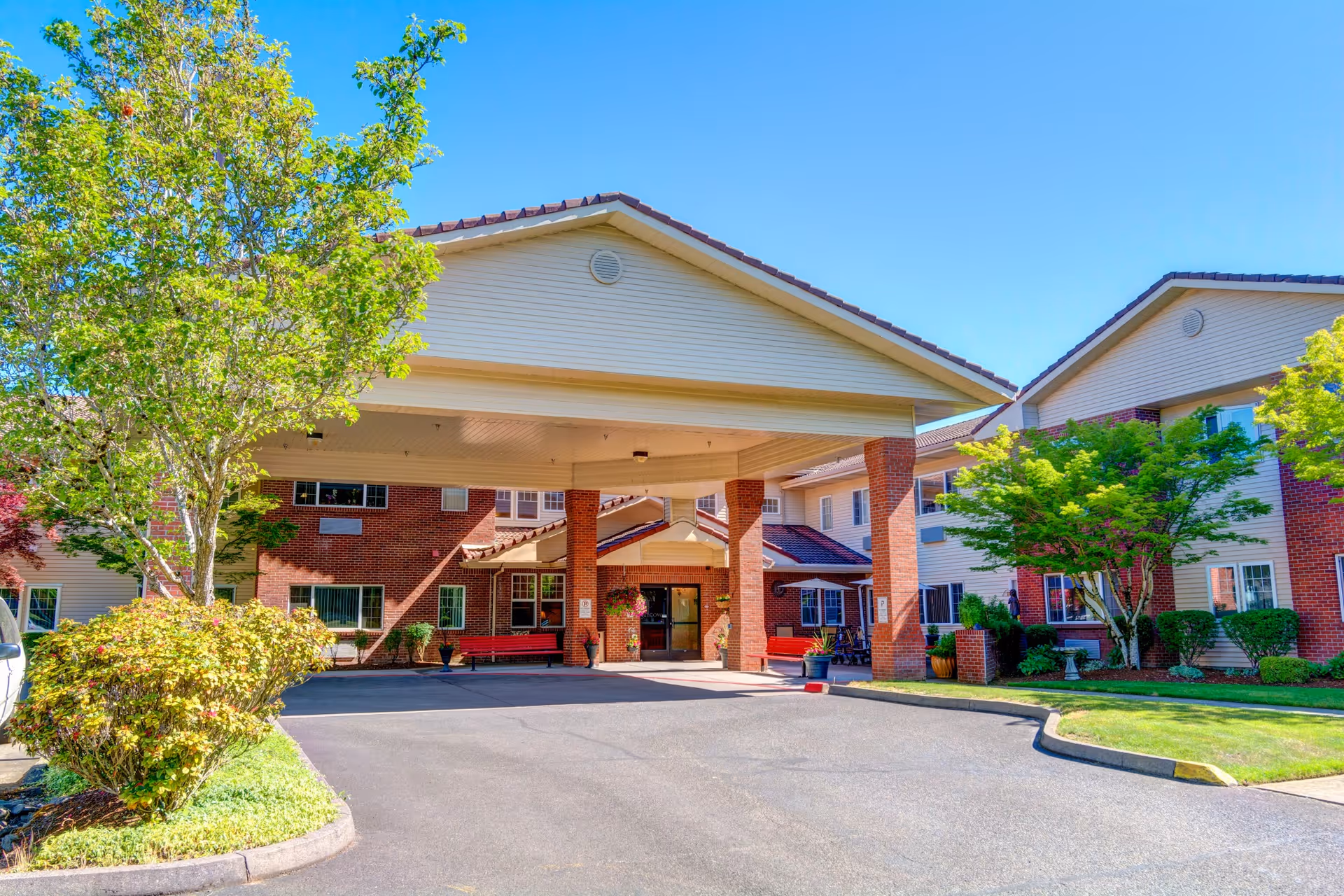 Front exterior view of Gilman Grove facility showing a covered entrance with brick pillars, surrounded by well-maintained landscaping including trees and shrubs under a clear blue sky.