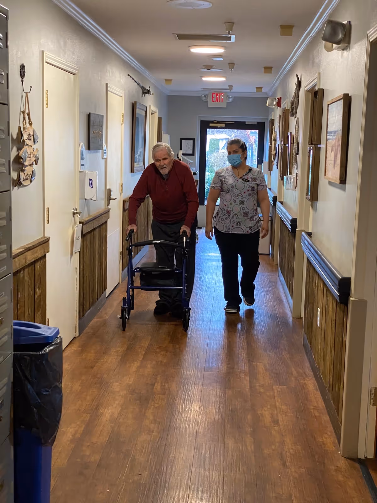 An elderly man using a walker is being assisted by a caregiver wearing a face mask as they walk down a well-lit hallway with wooden flooring and framed pictures on the walls.
