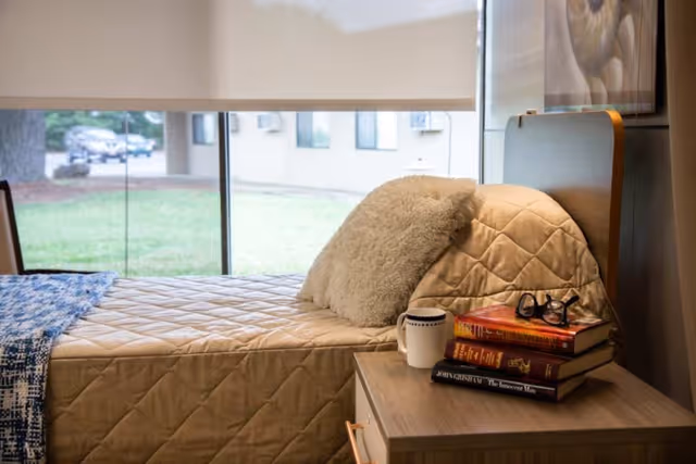 A neatly made single bed with a fluffy pillow and a bedside table holding stacked books, glasses and a mug next to a large window.