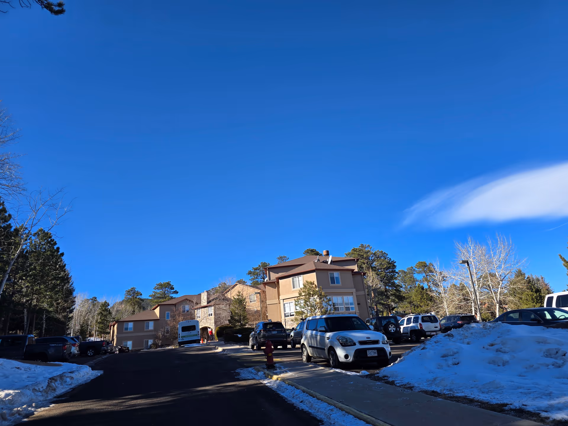 Exterior view of a multi-story assisted living facility building named Elk Run Assisted Living, surrounded by parked cars, trees, and patches of snow under a clear blue sky.