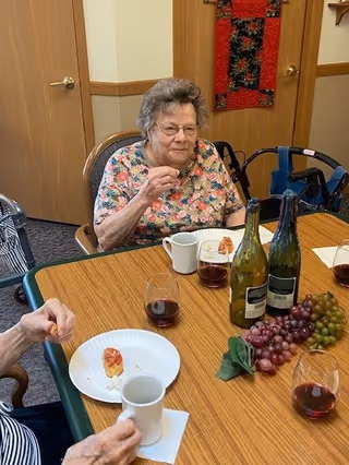 An elderly woman sitting at a table in a room, enjoying a small snack with a glass of red wine. The table has two bottles of wine, bunches of red and green grapes, and several glasses of red wine. Another person is partially visible holding a white mug and a small snack on a paper plate.