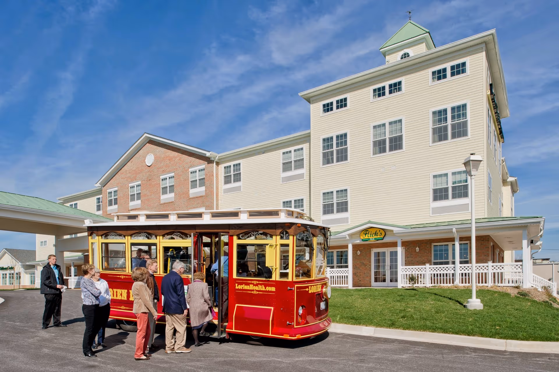 A red trolley with several people stopped in front of a multi-story senior living building under a blue sky.