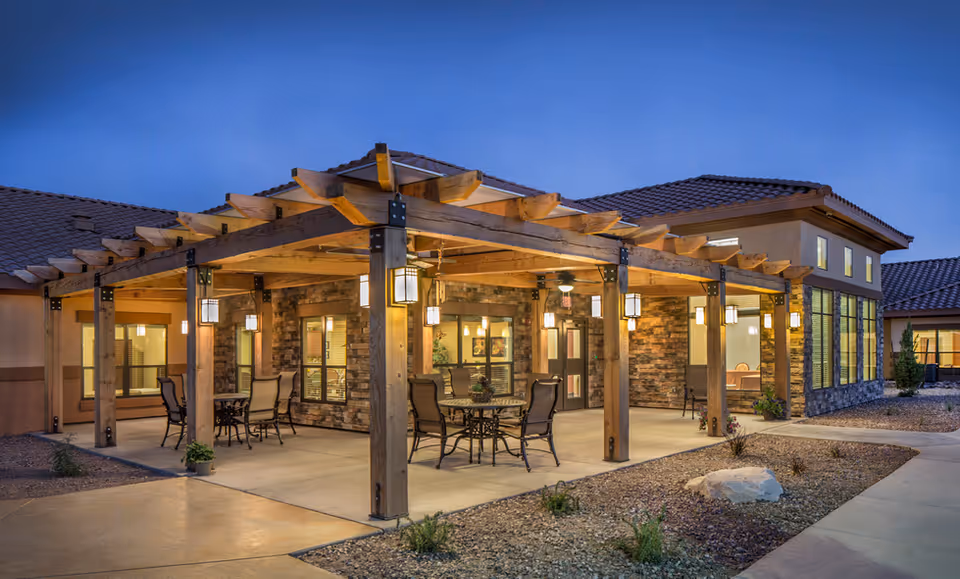 Covered outdoor patio with wooden pergola, tables and chairs outside a lit senior living building at dusk.