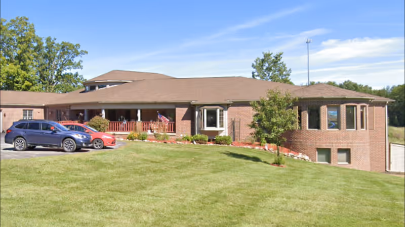 Single-story brick senior living facility with a front porch, grassy lawn, and parked cars.