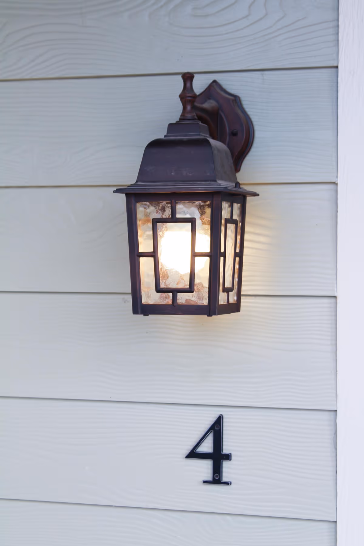 A lit decorative outdoor wall lantern mounted on light-colored siding above a black house number 4.