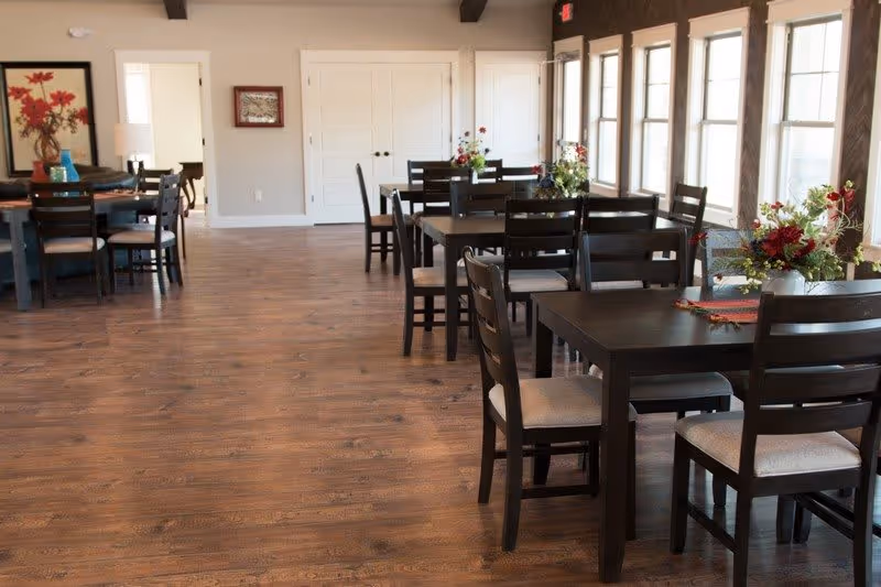 A bright dining room with multiple dark wooden tables and chairs arranged neatly. Each table has a floral centerpiece. Large windows on the right side allow natural light to fill the room, and the floor is made of polished wood.