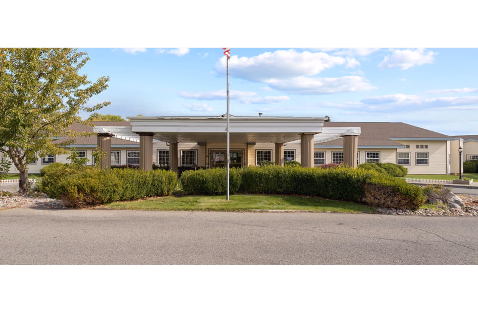Front entrance of a care center showing a covered porte-cochere with columns, a flagpole, and landscaped shrubs.