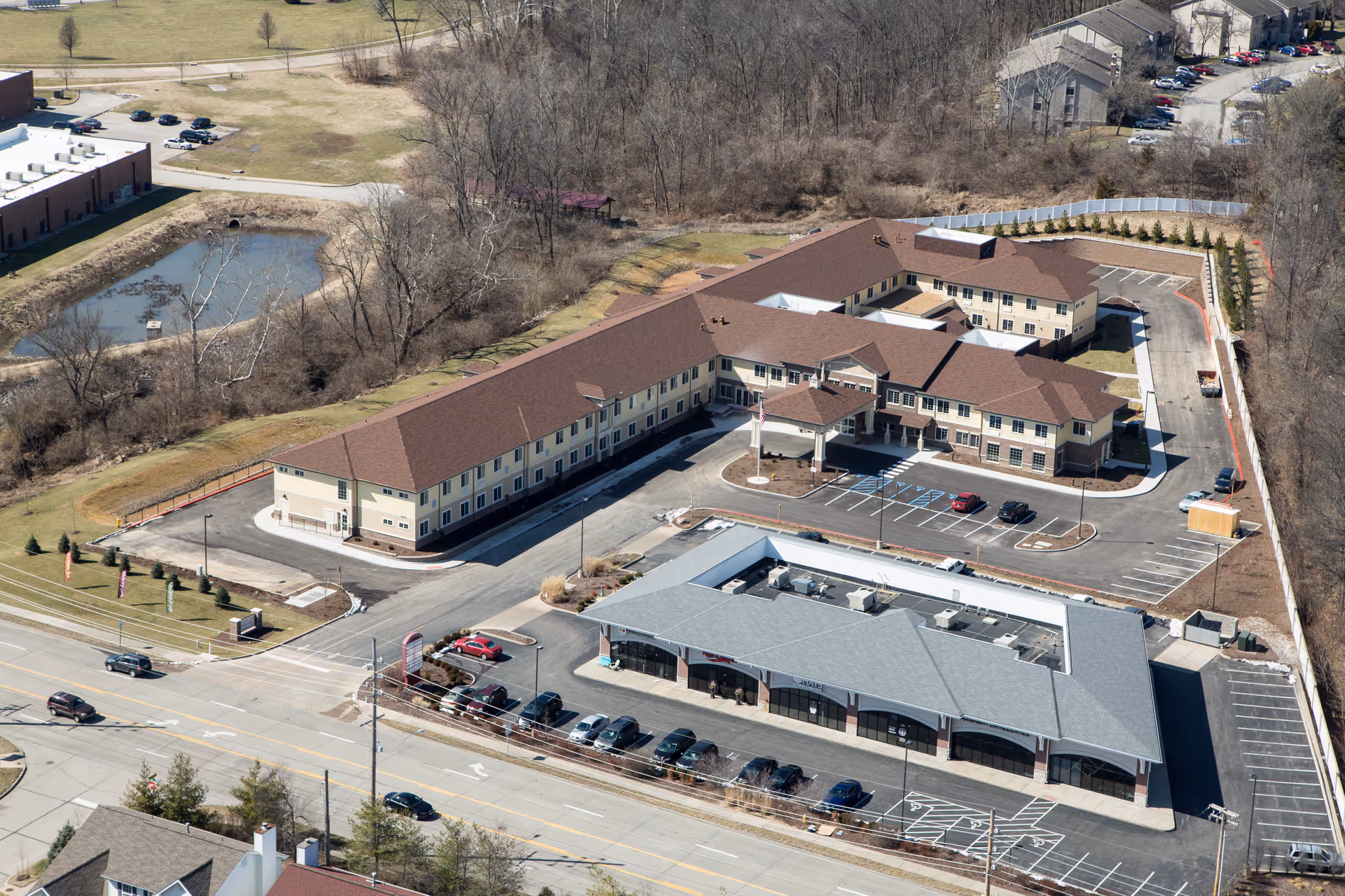 Aerial view of Dougherty Ferry Assisted Living and Memory Care facility showing a large two-story building with a brown roof and beige walls, surrounded by parking lots and adjacent commercial buildings. The area includes some trees, a small pond, and a road with cars in front of the facility.