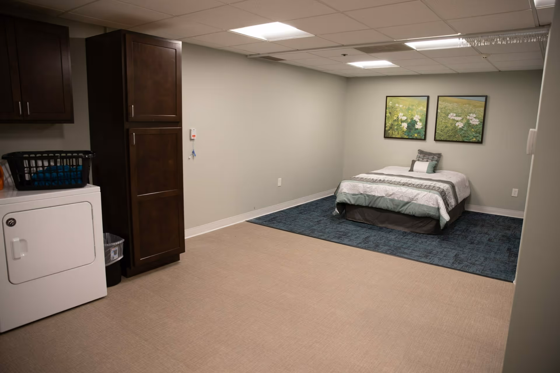 A simple bedroom with a bed placed on a blue rug against a light gray wall. Above the bed are two framed pictures of flowers in a field. To the left, there is a white dryer with a laundry basket on top and dark wooden cabinets above and beside it. The floor is covered with beige carpet, and the ceiling has fluorescent lighting.