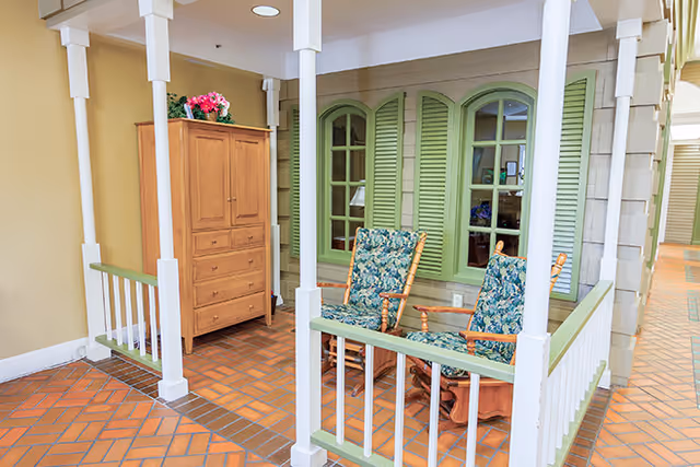 Indoor seating area with two wooden rocking chairs featuring floral cushions, a small wooden table between them, green window shutters on the wall behind, and a wooden cabinet with drawers and a flower pot on top. The floor is tiled with a reddish-brown pattern and white pillars with green railings surround the seating area.