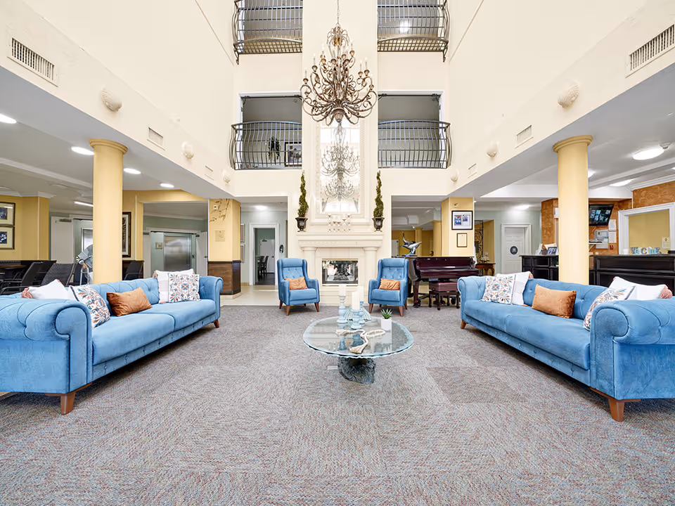 Spacious and bright living room area with two blue sofas and two blue armchairs arranged around a glass coffee table with decorative items. The room features a high ceiling with a large chandelier, a fireplace with a mirror above it, and yellow columns. There is a piano and additional seating in the background, with open balconies on the upper level.