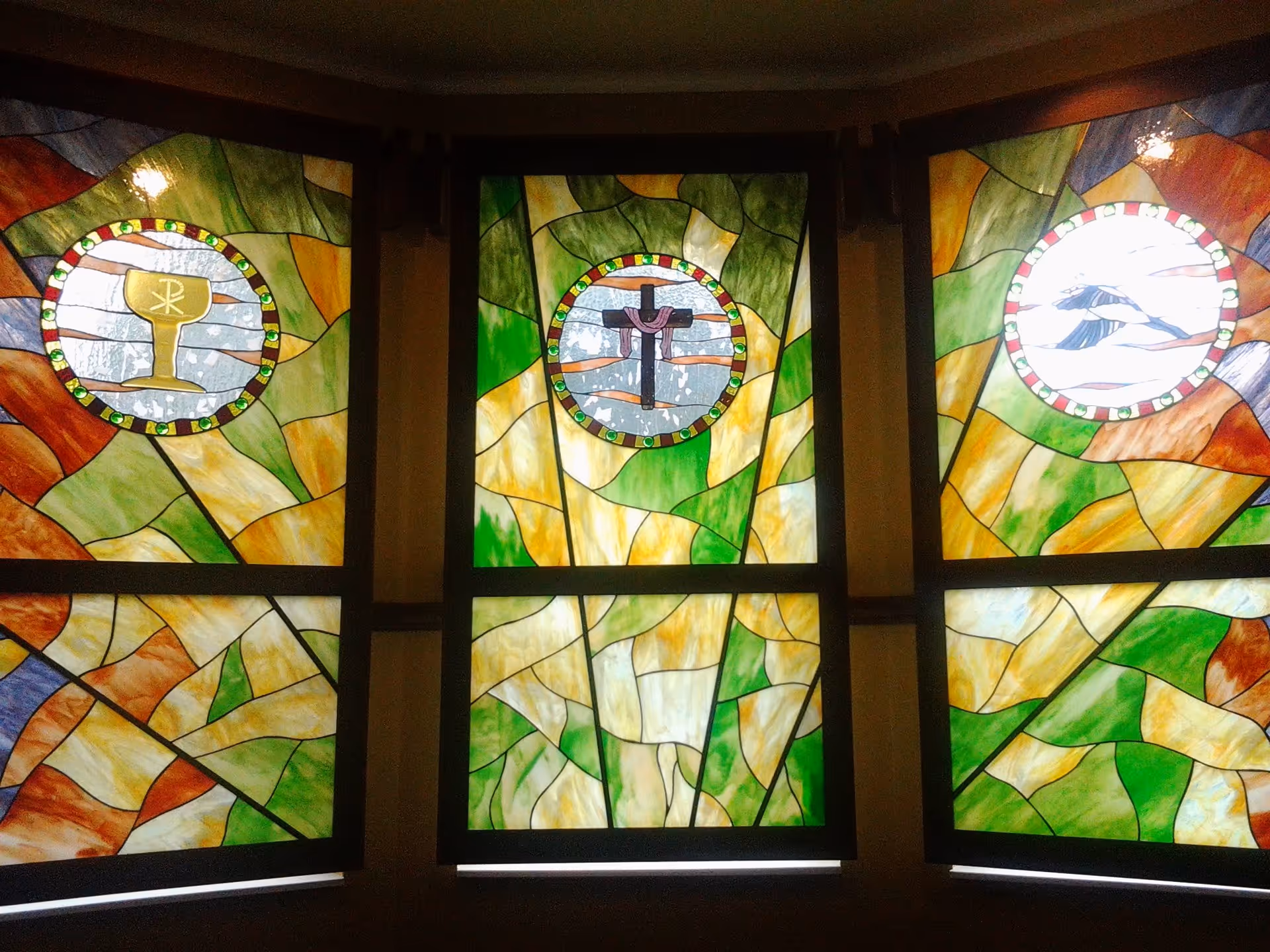 Three stained glass windows with colorful abstract patterns in green, yellow, orange, and blue. Each window has a circular emblem in the center: the left window shows a golden chalice, the middle window depicts a cross with a cloth draped over it, and the right window features a bird in flight.