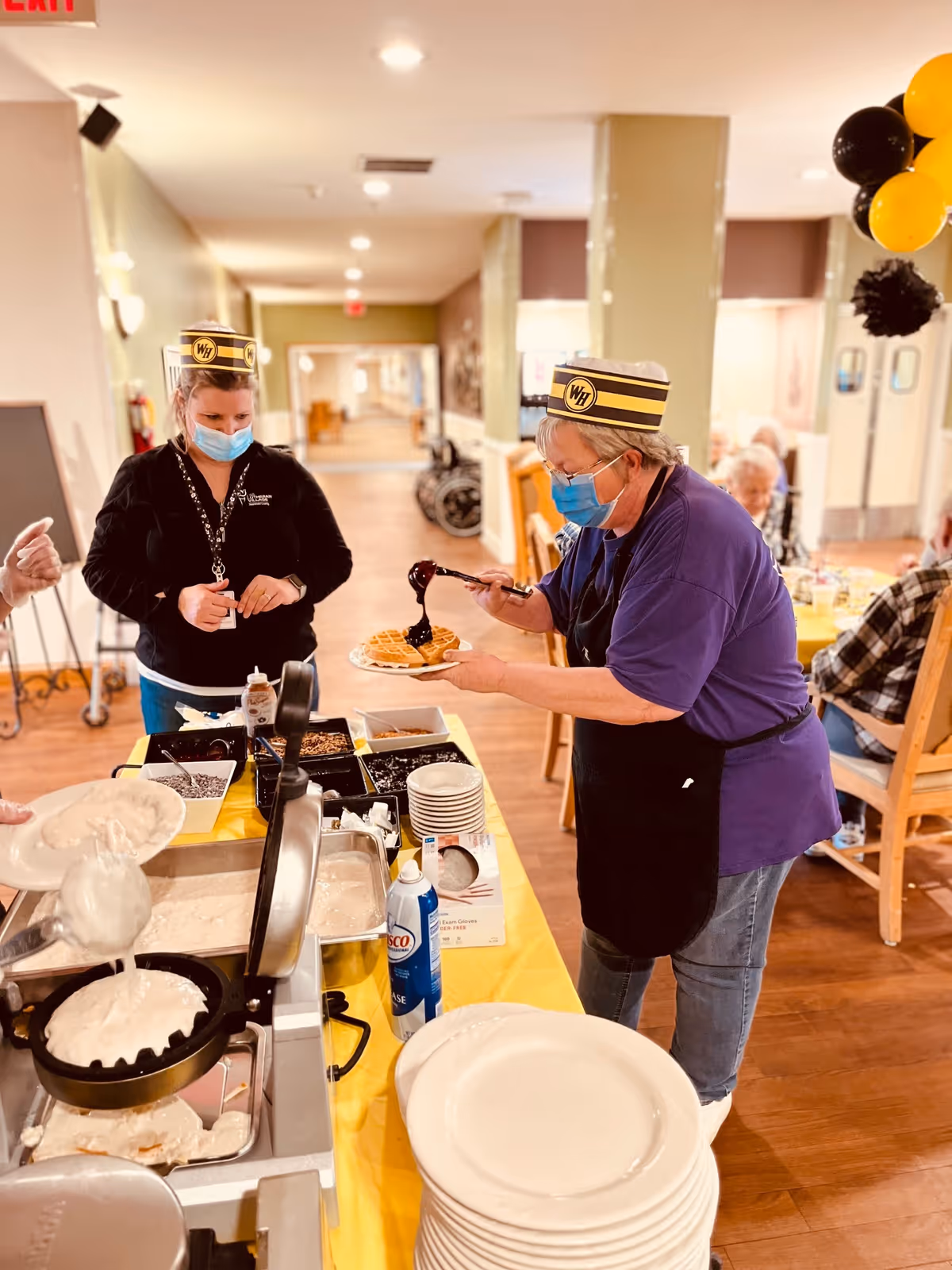 Staff wearing masks serve waffles at a buffet table in a senior living dining room.