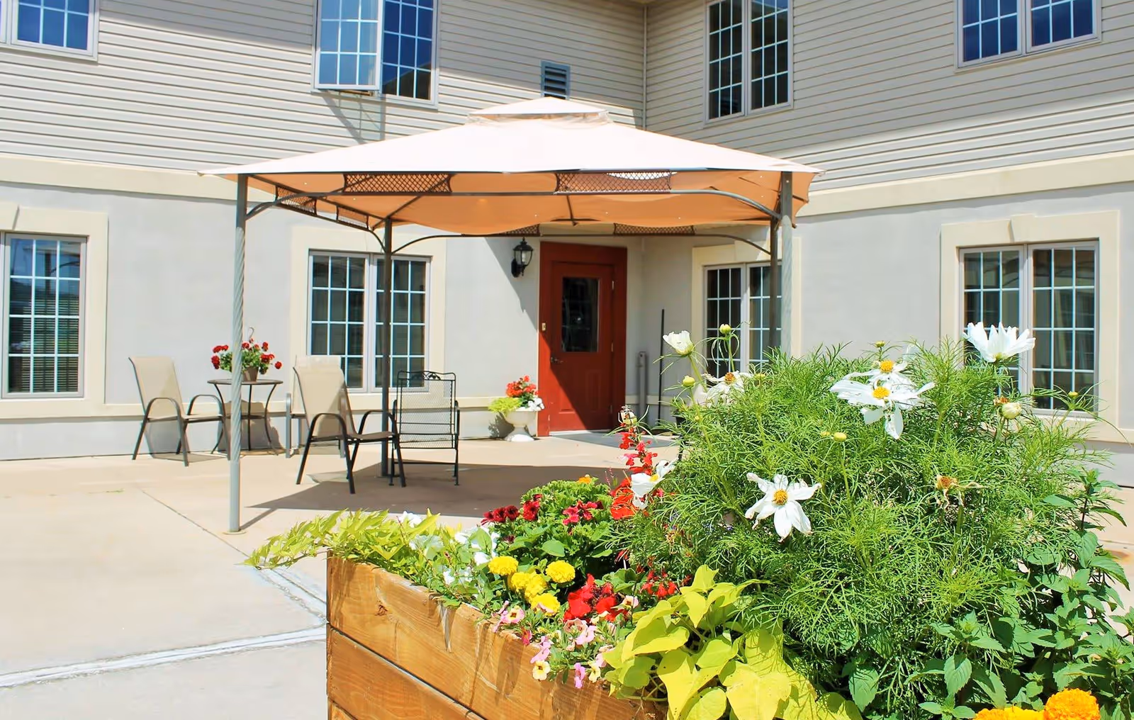 Outdoor patio area at Mountain Terrace Senior Living with a large beige canopy providing shade over several chairs and a small table. The patio is adjacent to a building with multiple windows and a red door. In the foreground, there is a wooden planter box filled with various colorful flowers and green plants.