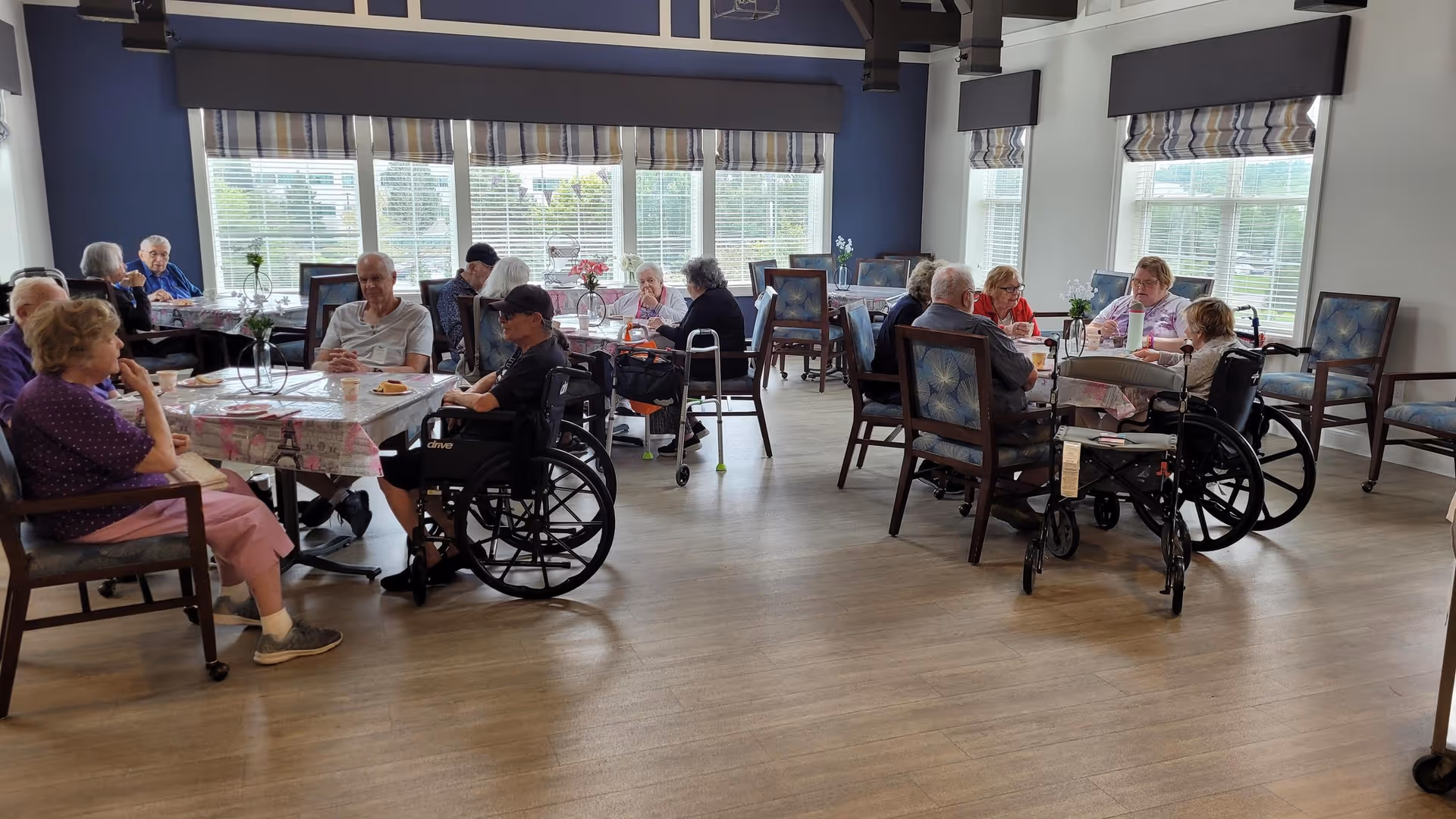 A group of elderly people sitting around tables in a bright dining room with large windows and wooden flooring. Some individuals are in wheelchairs, and the tables are covered with patterned tablecloths and small flower vases.