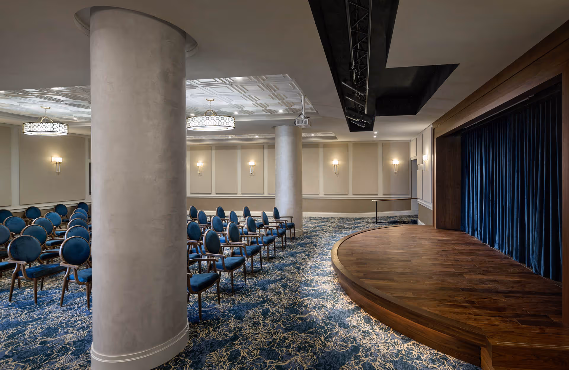 Well-lit meeting room with rows of blue upholstered chairs, decorative columns, and a wooden stage with blue curtains.