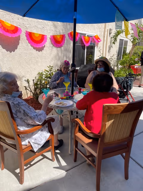Four elderly women sitting around a round outdoor table under a blue umbrella, enjoying drinks and snacks. The setting is a sunny patio area decorated with colorful pink and orange paper garlands on the wall. Some of the women are wearing face masks, and there are plants and windows in the background.