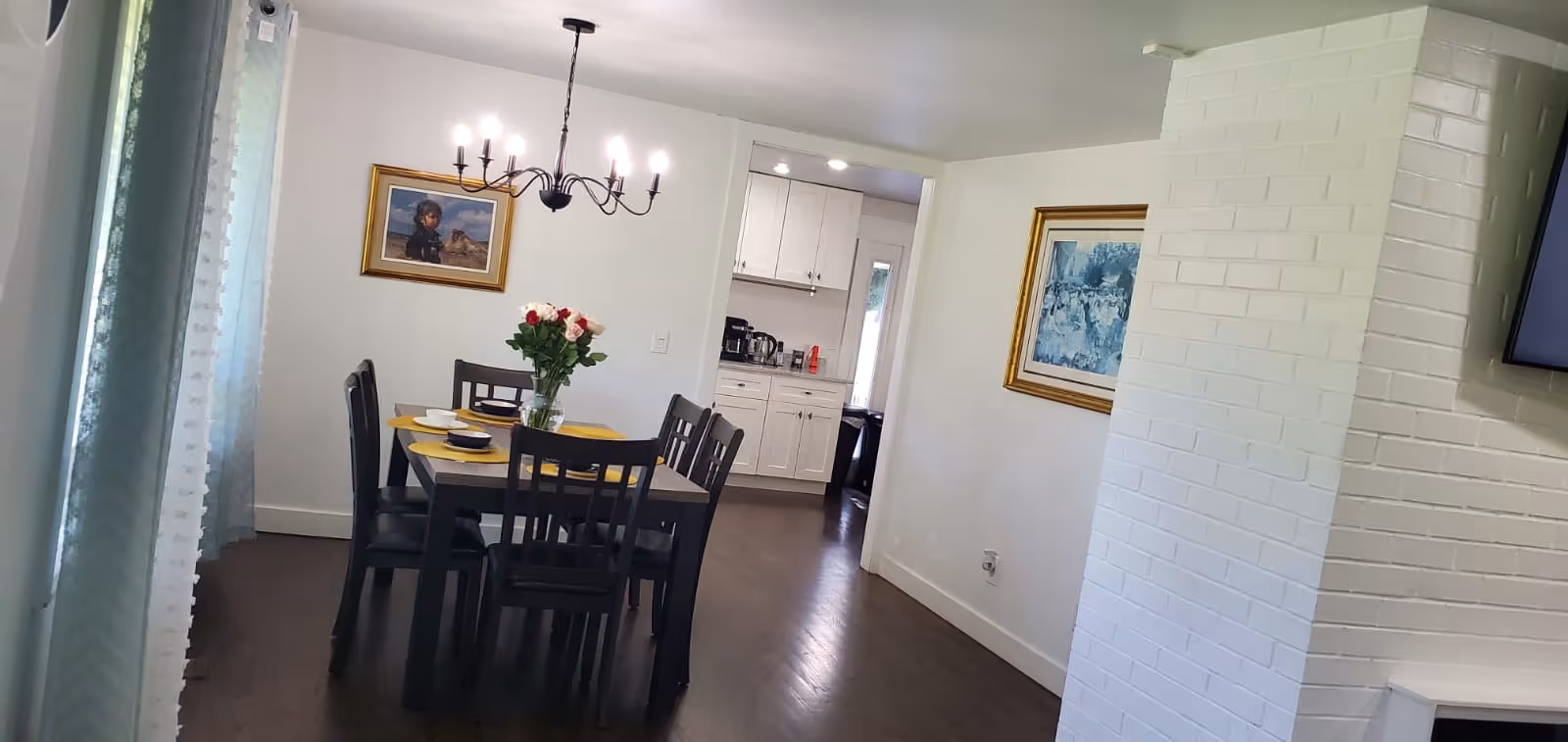 A dining room with a rectangular wooden table set for six with yellow placemats and a vase of flowers in the center. There is a chandelier with lit bulbs hanging above the table. The room has white walls with two framed paintings and dark wooden flooring. A doorway leads to a kitchen with white cabinets and appliances visible.