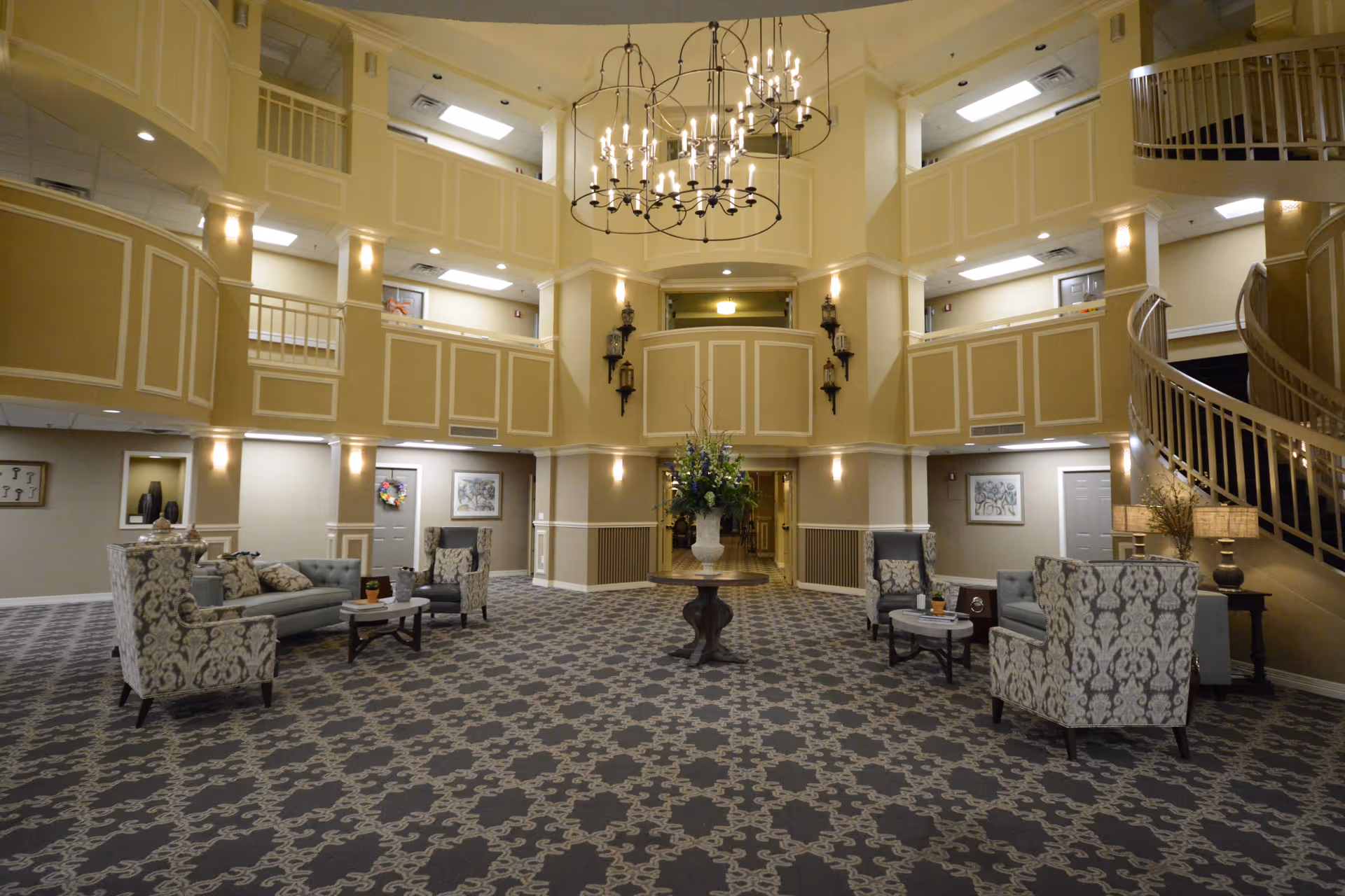 Spacious and elegant common area in a senior living facility with patterned carpet, multiple seating arrangements including sofas and armchairs, a central table with a floral arrangement, high ceilings with chandeliers, and a curved staircase leading to the upper floors.