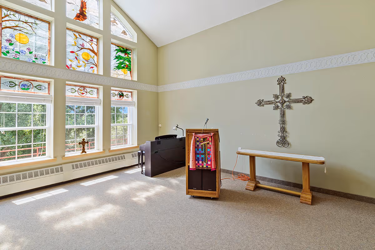 A bright room with large windows featuring colorful stained glass panels at the top. The room has a podium with a microphone and a colorful cloth draped over it, a wooden bench, and a decorative metal cross mounted on the wall. Sunlight streams through the windows, casting light patterns on the carpeted floor.