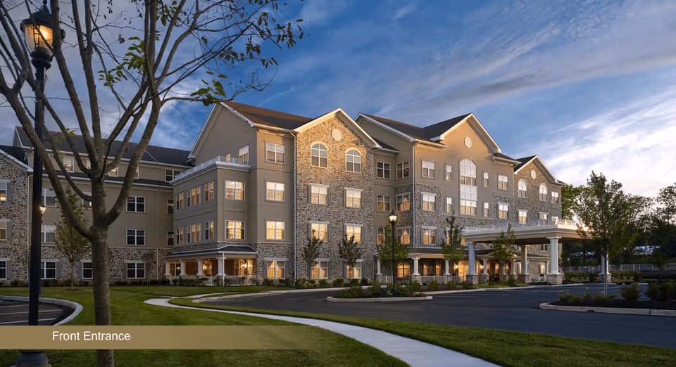 Four-story senior living building front entrance with lit windows, covered porte-cochère, driveway and landscaped grounds at dusk.