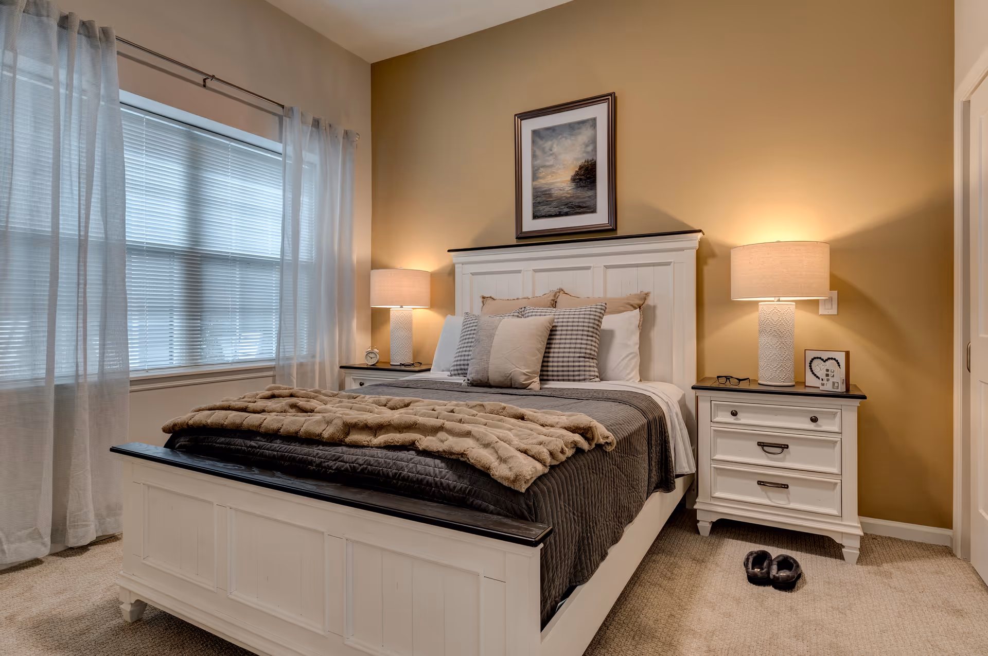 A cozy bedroom with a white wooden bed frame and matching nightstands on either side. The bed is made with gray and beige bedding, multiple pillows, and a soft throw blanket. Two lamps with beige shades sit on the nightstands, illuminating the room. A framed painting hangs above the headboard. Sheer white curtains cover a large window on the left side, and a pair of slippers is placed on the carpeted floor near the right nightstand.
