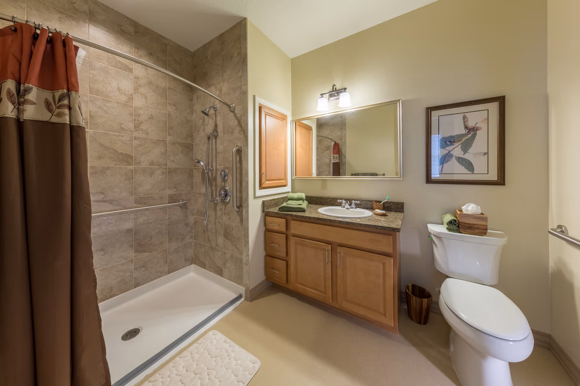A clean bathroom featuring a walk-in shower with beige tiled walls and a brown and red shower curtain. There is a wooden vanity with a granite countertop, a sink, and a large mirror above it. Next to the vanity is a white toilet with a tissue box and green towel on top. A framed picture hangs on the wall above the toilet, and a small trash bin is placed beside it.