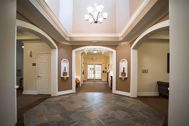 Interior view of a senior living facility lobby area with a tiled floor, beige walls, white trim, and a chandelier hanging from a recessed ceiling. There are archways leading to other rooms and a glass door at the far end letting in natural light.
