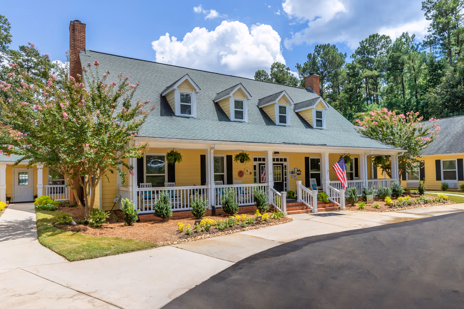 Front exterior of a yellow senior living building with a covered porch, dormer windows, landscaping, and an American flag.