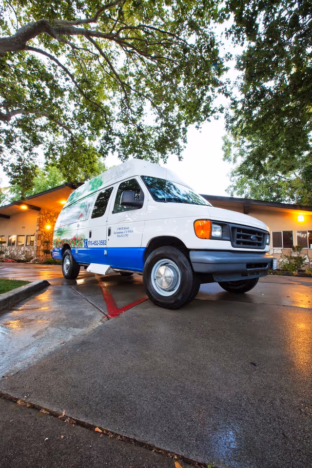 A white and blue shuttle van parked on a wet driveway outside a building surrounded by trees with green leaves. The building has stone and beige walls with windows and exterior lights on. The scene appears to be taken during early evening or late afternoon.