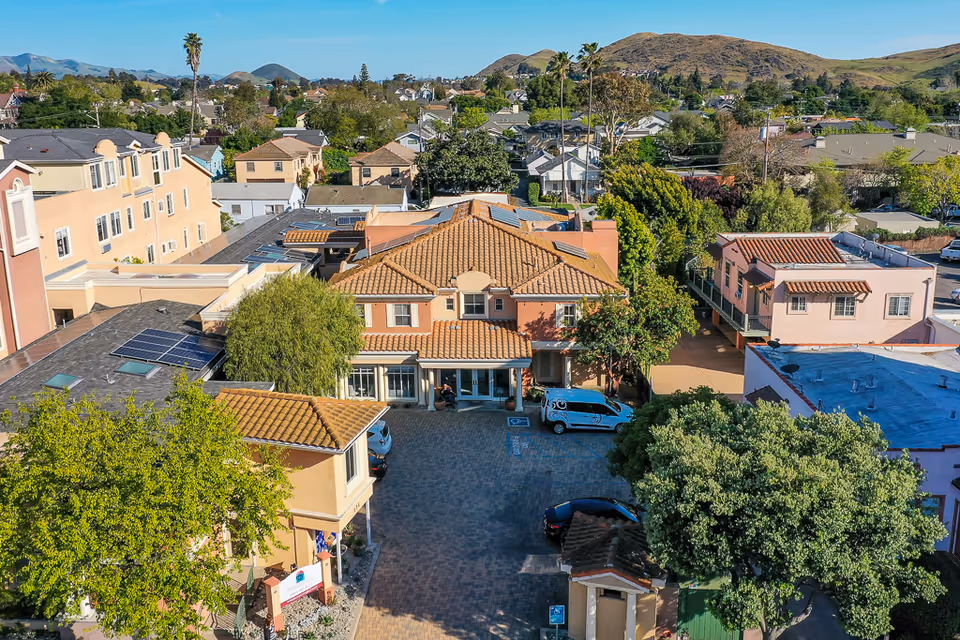 Aerial view of a senior living facility with terracotta-tiled roofs, a paved courtyard with parked vehicles, surrounding houses and distant hills.