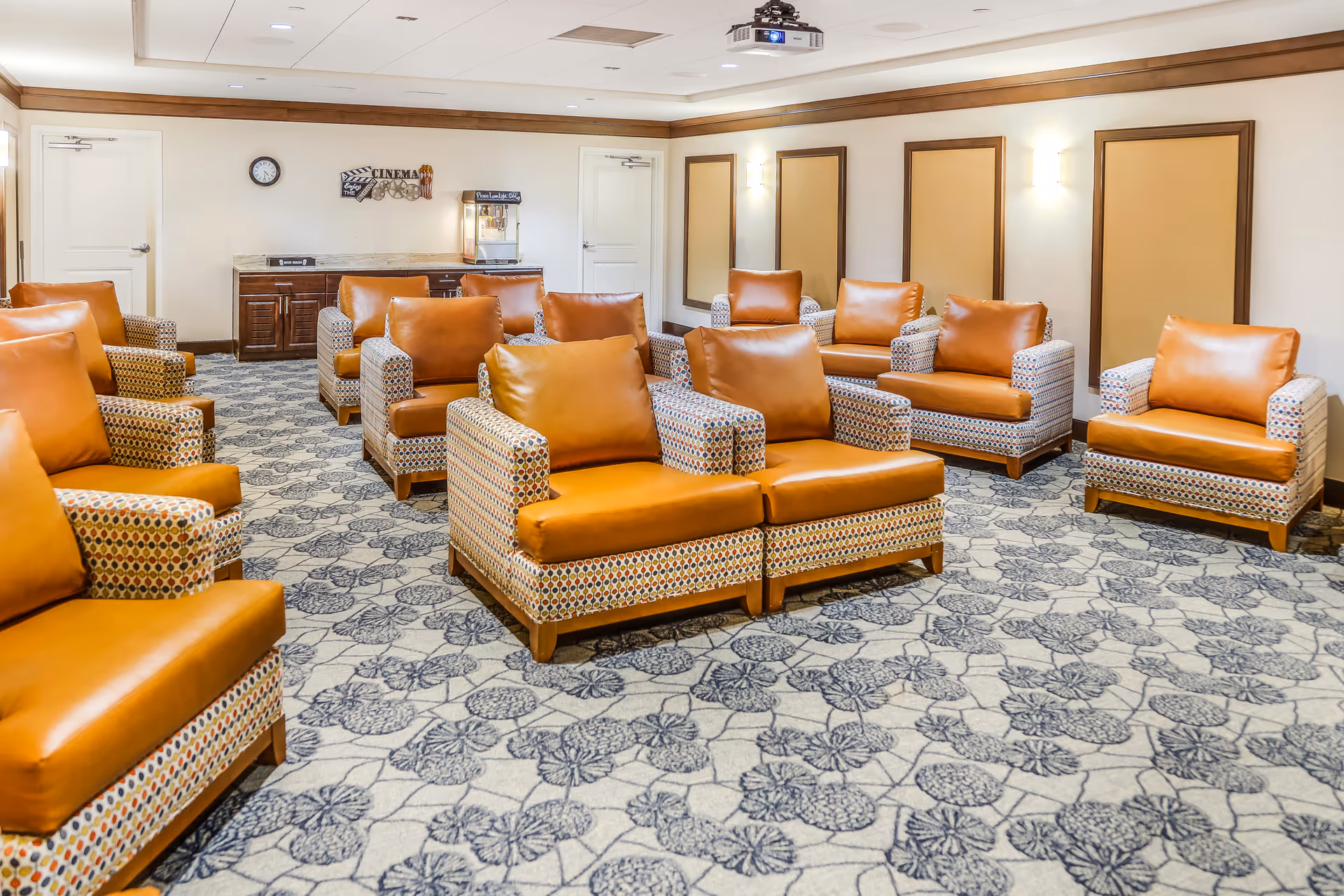 A cozy room with multiple patterned armchairs featuring brown leather cushions arranged in rows on a floral carpet. The room has beige walls with wooden trim, wall-mounted lights, a clock, a popcorn machine, and a cinema-themed wall decoration.