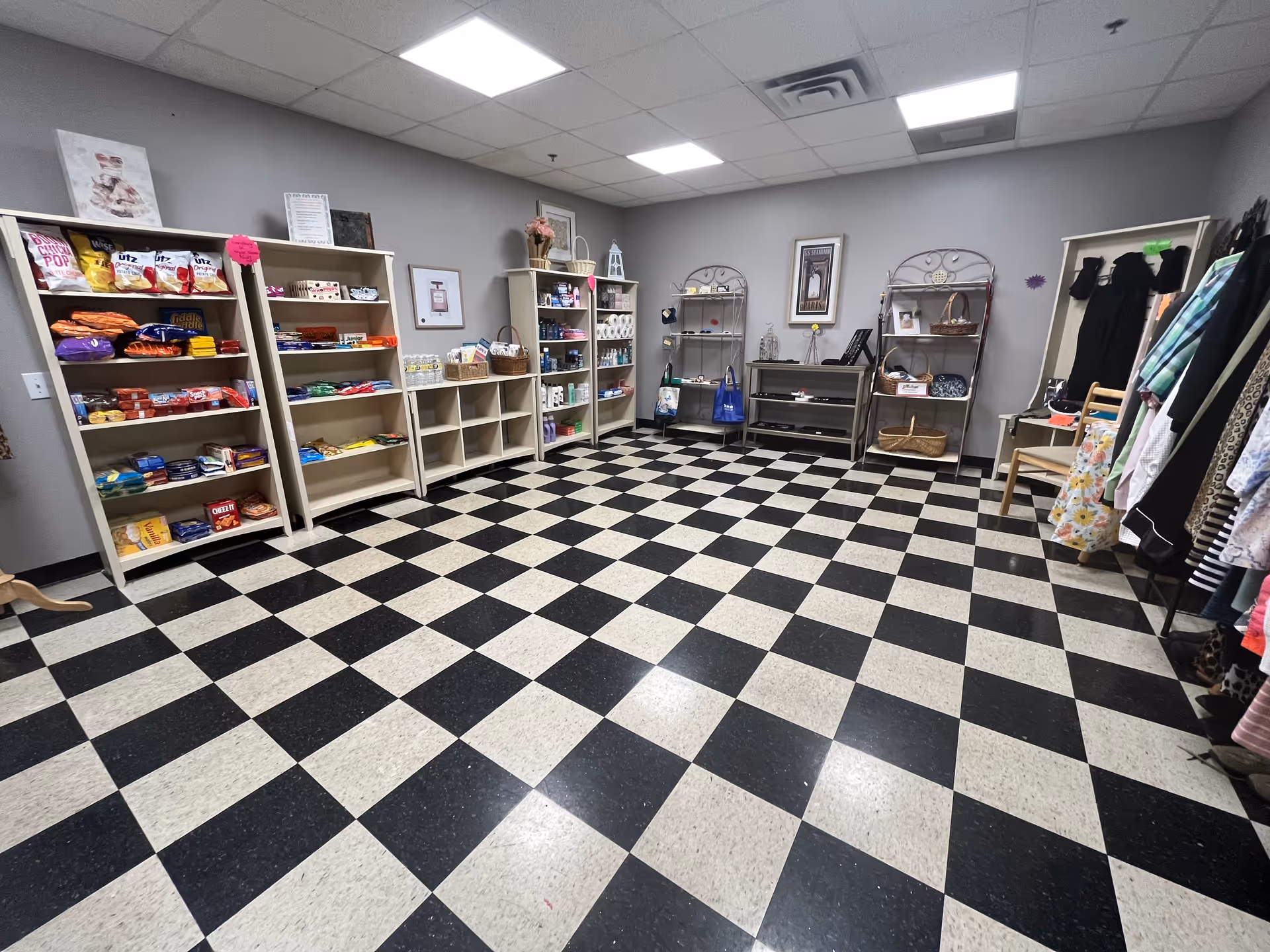 Room with black and white checkered floor and gray walls, featuring shelves stocked with snacks, household items, and clothing racks with various garments.