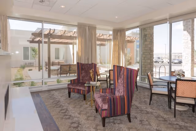 A bright and spacious common area with large windows showing an outdoor patio. The room features two striped upholstered armchairs with a small side table between them, a round table with four chairs, and light-colored curtains. The floor is carpeted with a patterned design.