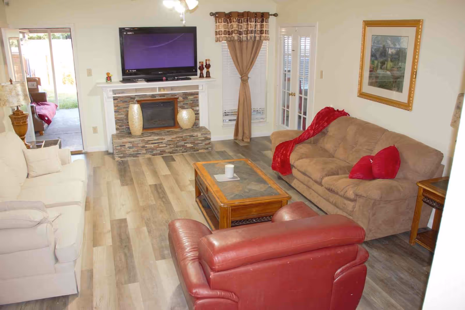 A cozy living room with a beige couch adorned with red pillows and a red throw blanket, a red leather armchair, and a cream-colored sofa. A wooden coffee table with a candle sits in the center on a wood-patterned floor. A flat-screen TV is mounted above a fireplace with decorative vases. The room has a window with brown curtains and a framed picture on the wall. French doors and a sliding door lead to other areas.