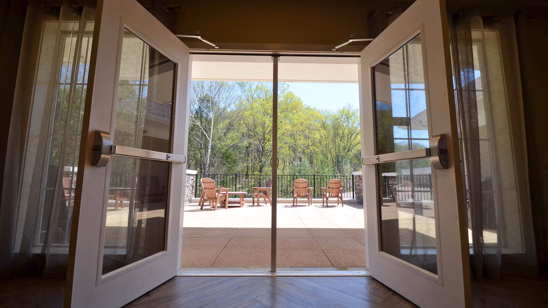 View through open double glass doors leading from an interior room to an outdoor patio area with several wooden chairs and tables. Beyond the patio, there are green trees and a clear sky.