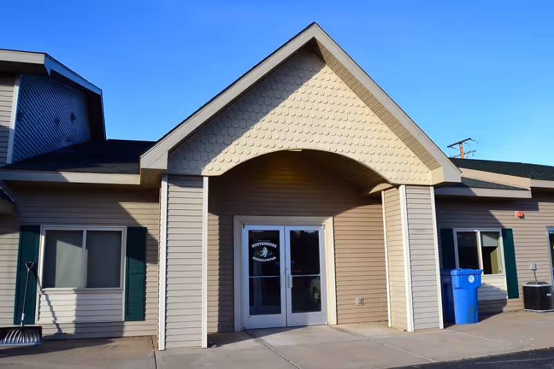 Exterior front entrance of a single-story building with beige siding and a peaked roof. The entrance has double glass doors with a logo on them, flanked by windows with green shutters. There is a blue recycling bin and a snow shovel near the building.