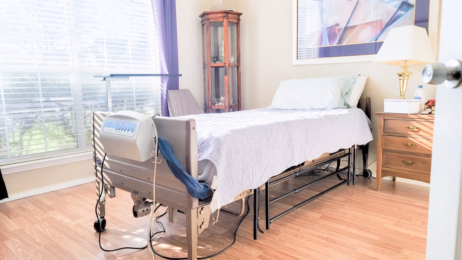 A bright room with a hospital-style bed covered with a light purple quilt. The bed has control buttons on the footboard and is positioned near a large window with blinds and purple curtains. There is a wooden nightstand with a lamp and some items on it, a wooden corner cabinet with glass doors, and a framed picture on the wall.