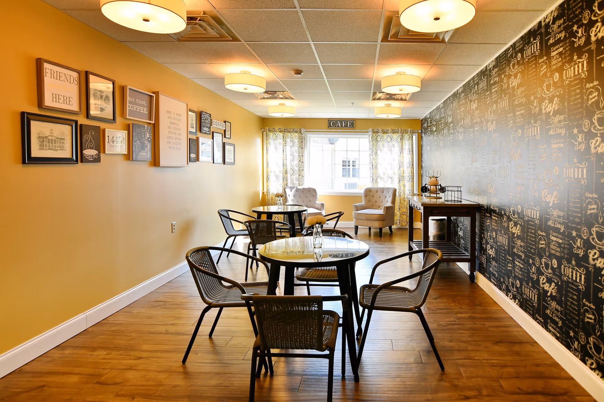 A cozy cafe-style room with wooden flooring, round tables with glass tops, and black metal chairs. The left wall is painted yellow and decorated with framed pictures and quotes. The right wall features black wallpaper with gold cafe-themed text. At the far end, two beige armchairs are placed near a window with patterned curtains, and a small table with coffee-related items is against the right wall.