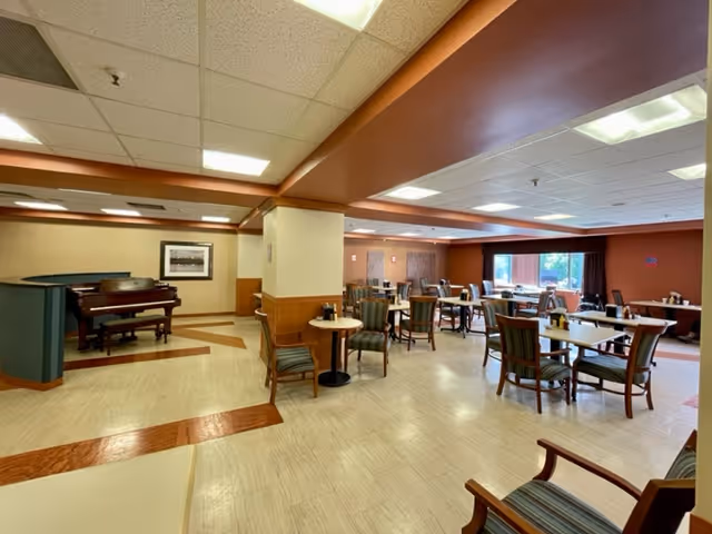A spacious dining room with multiple tables and chairs arranged neatly. The room has a beige and brown color scheme with a piano in the corner and large windows allowing natural light to enter.