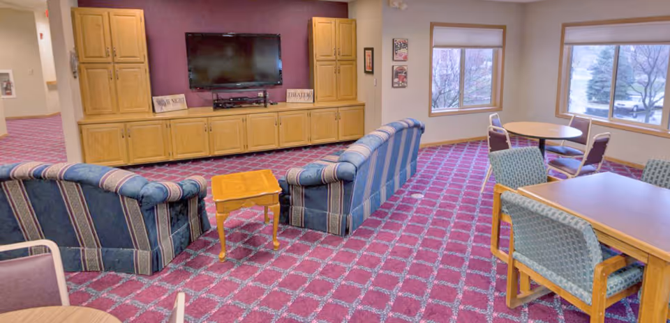 A senior living facility common area with patterned purple carpet, two blue striped sofas facing a wall-mounted TV on a wooden cabinet, and several tables with chairs near large windows showing trees outside.