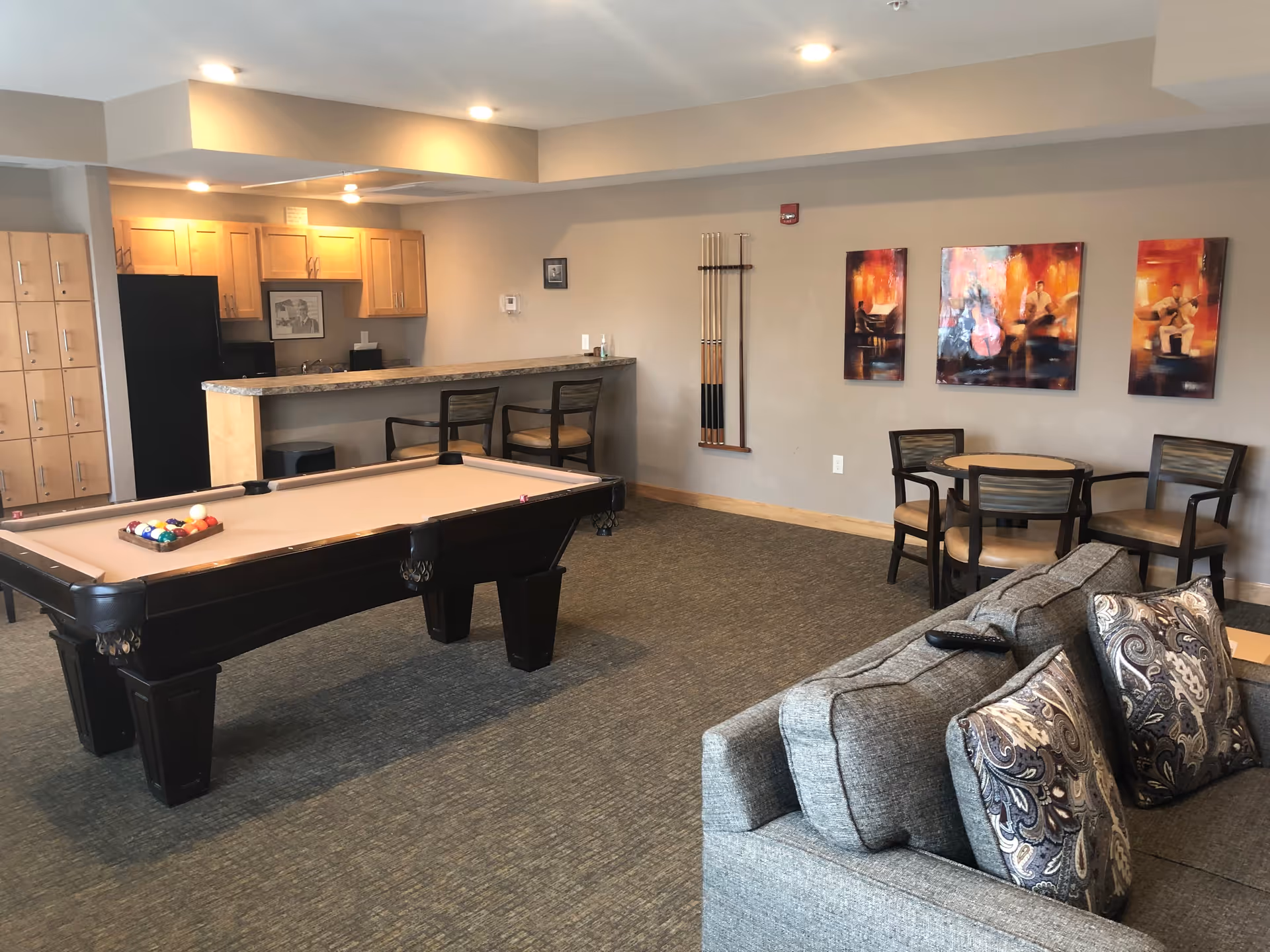 A senior community common area featuring a pool table with balls arranged for a game, a gray couch with patterned pillows, a small round table with four chairs, and a kitchenette with wooden cabinets and a countertop with two bar stools. The walls are decorated with three colorful paintings and a rack holding pool cues.