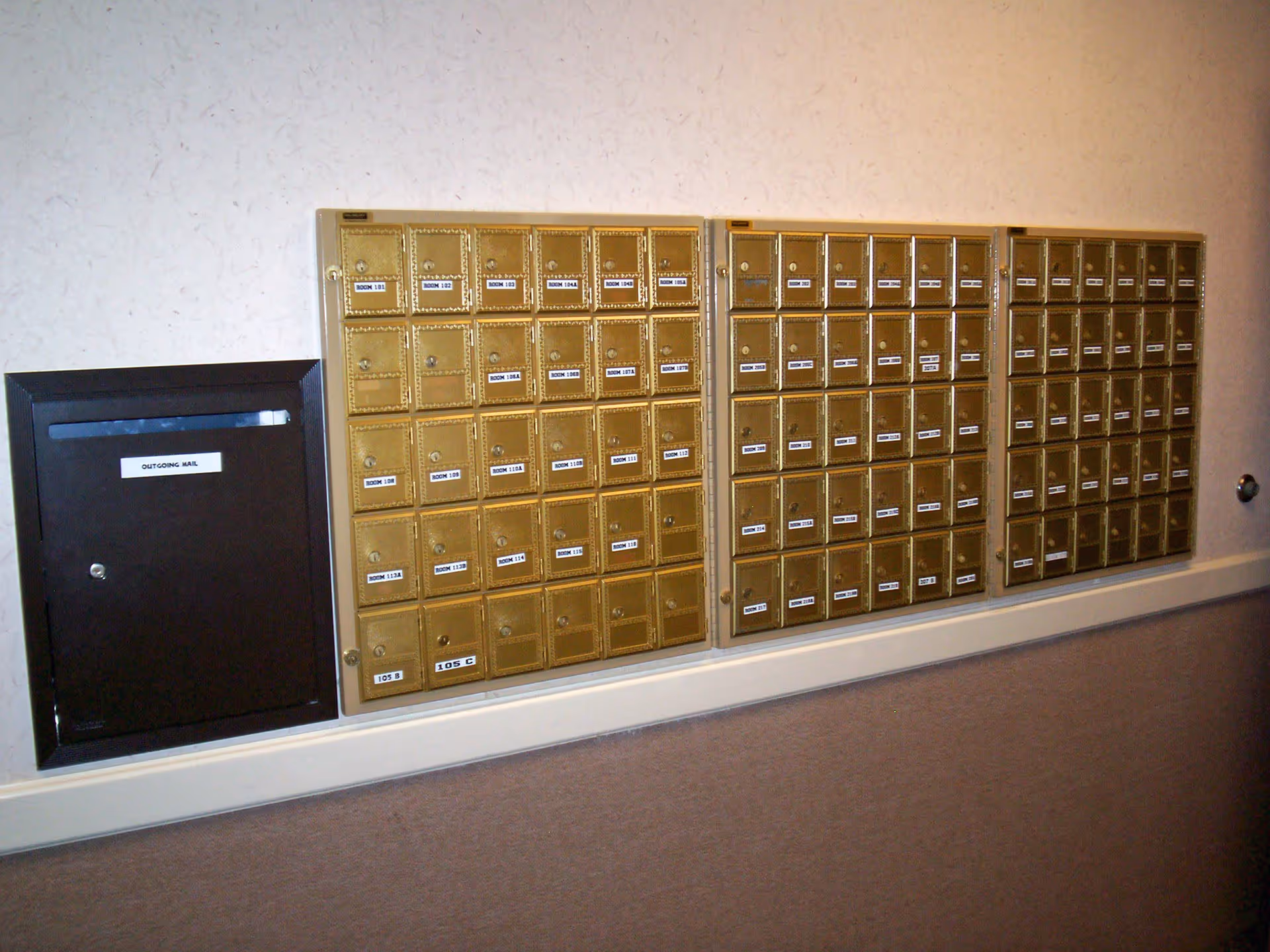 A wall-mounted set of multiple small brass mailboxes labeled with room numbers, next to a black outgoing mail slot, all installed on a beige wall.