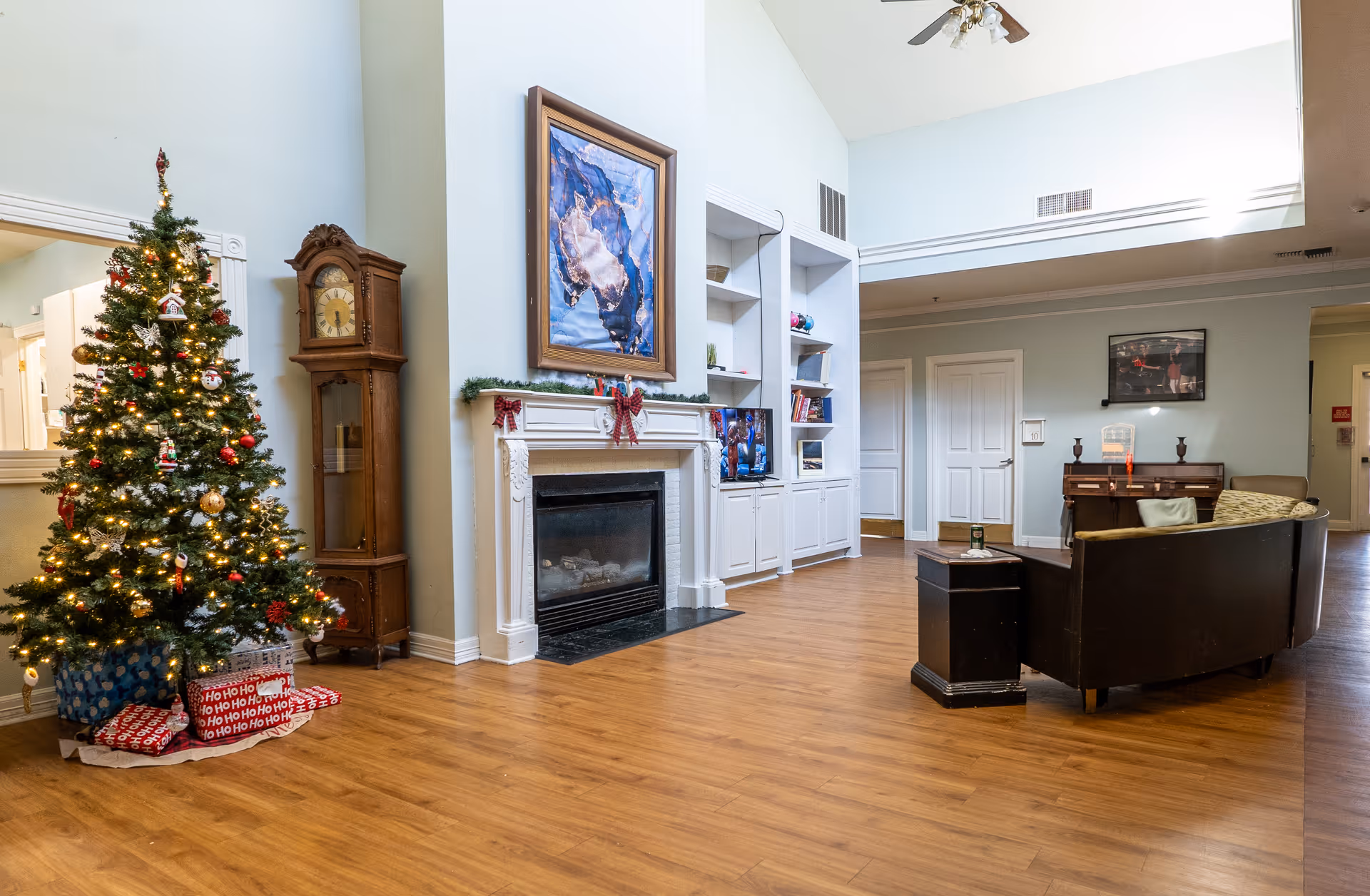 A spacious living room area decorated for Christmas with a lit Christmas tree and wrapped presents underneath. The room features a wooden grandfather clock, a white fireplace with holiday garland and red bows, built-in white shelves with books and decorations, a wall-mounted TV, a dark curved sofa with cushions, a small black side table, and a piano against the far wall. The floor is wooden, and the walls are painted light blue with white trim.