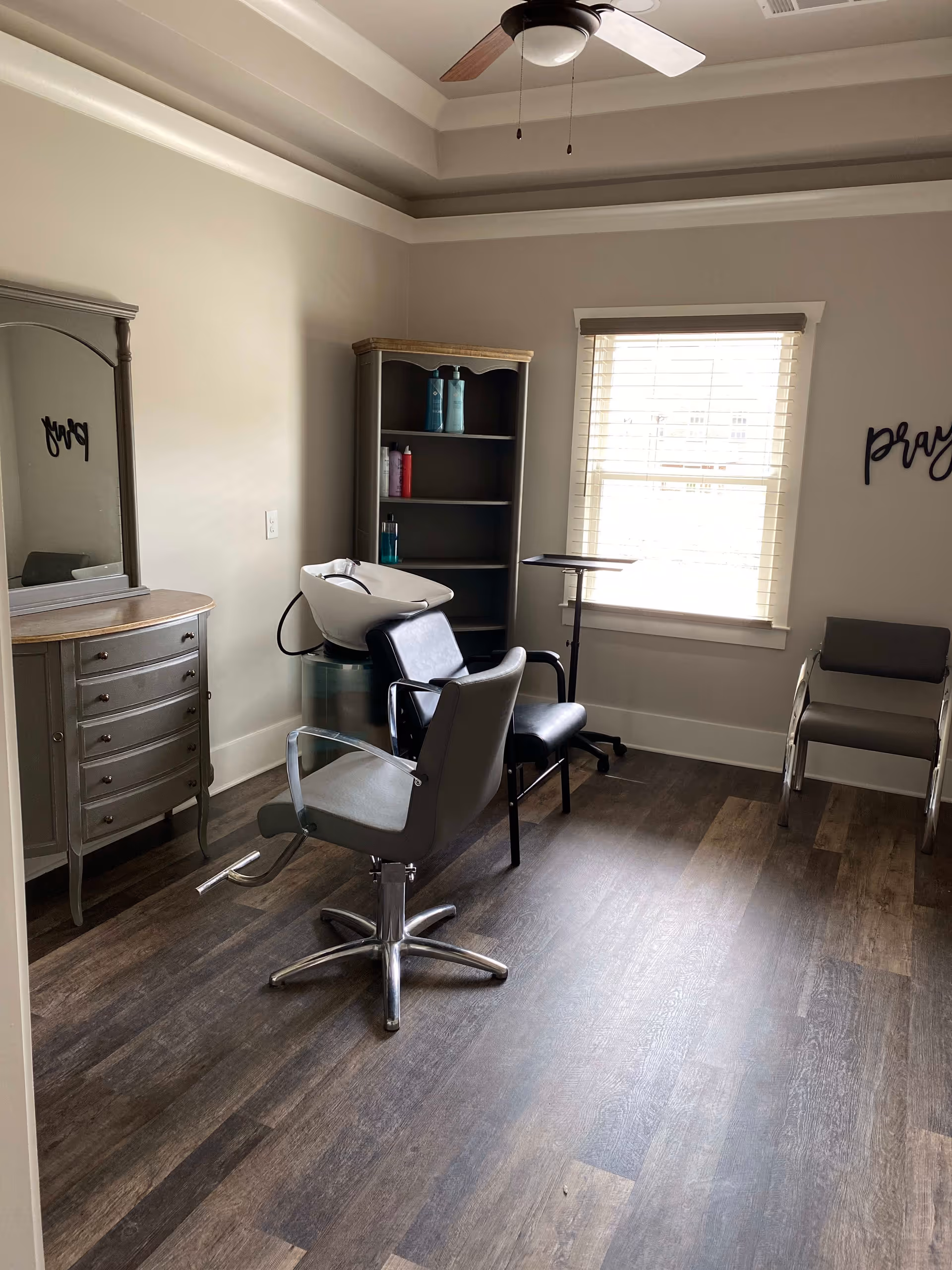 Small salon room with styling chairs, a shampoo sink, shelving with hair products, a mirror and a window.