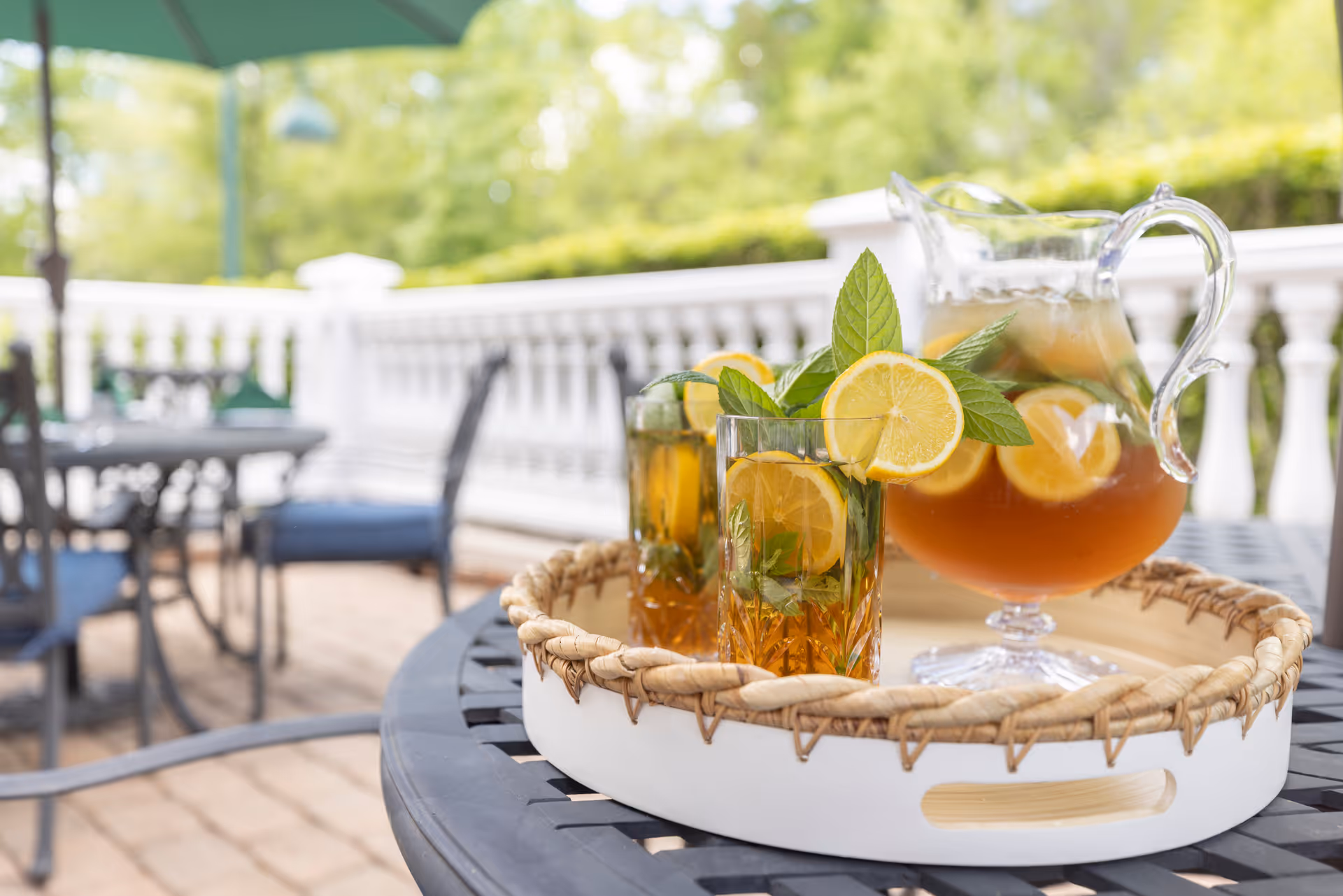 A close-up view of a tray with a glass pitcher and two glasses of iced tea garnished with lemon slices and fresh mint leaves, placed on a black outdoor table on a patio with chairs and a white railing in the background.