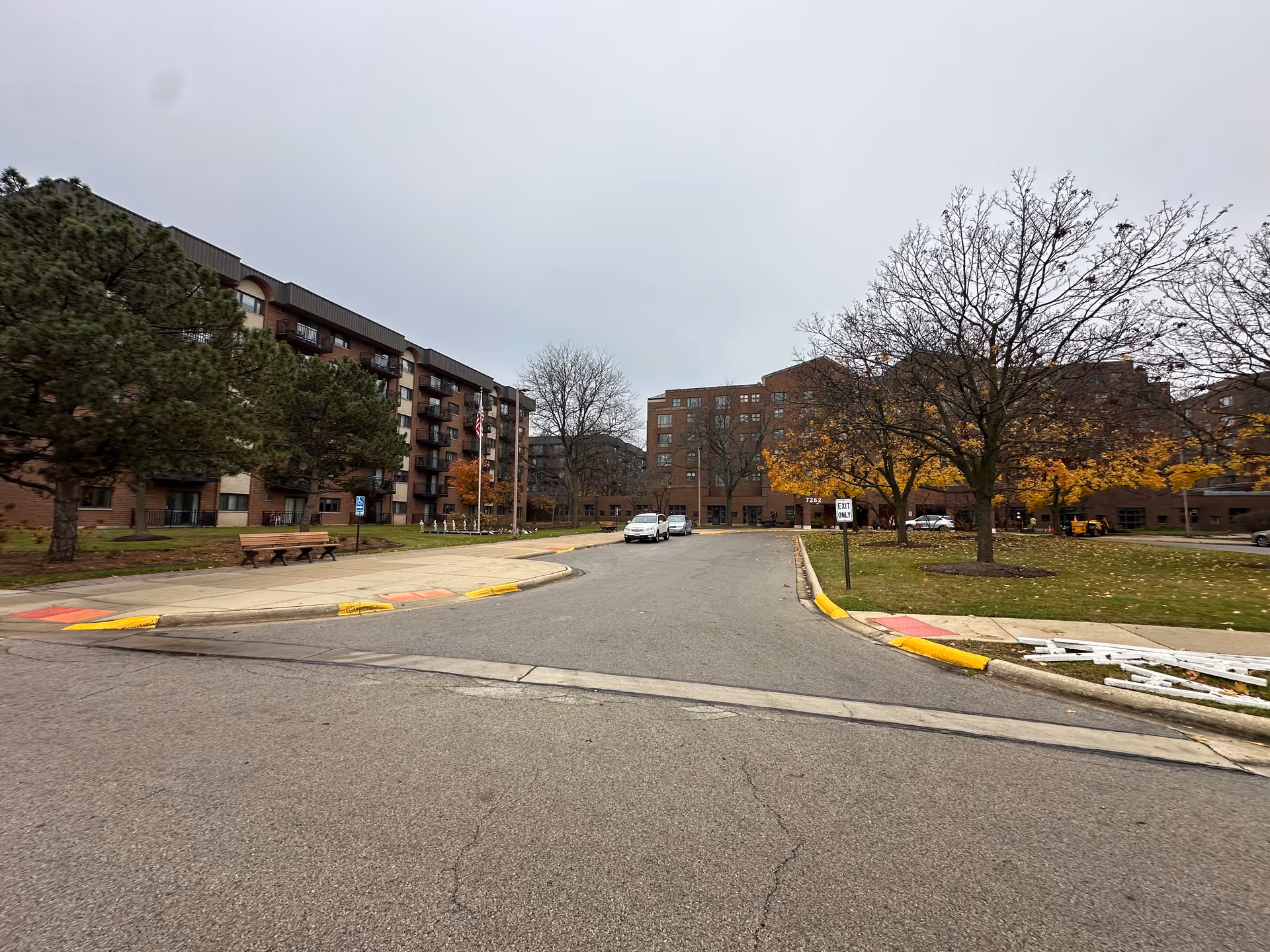 Exterior view of Ascension Living Resurrection Village showing a driveway leading to the entrance of a multi-story brick building with several windows and balconies. There are trees with autumn foliage, a few parked cars, benches on the sidewalk, and a cloudy sky overhead.