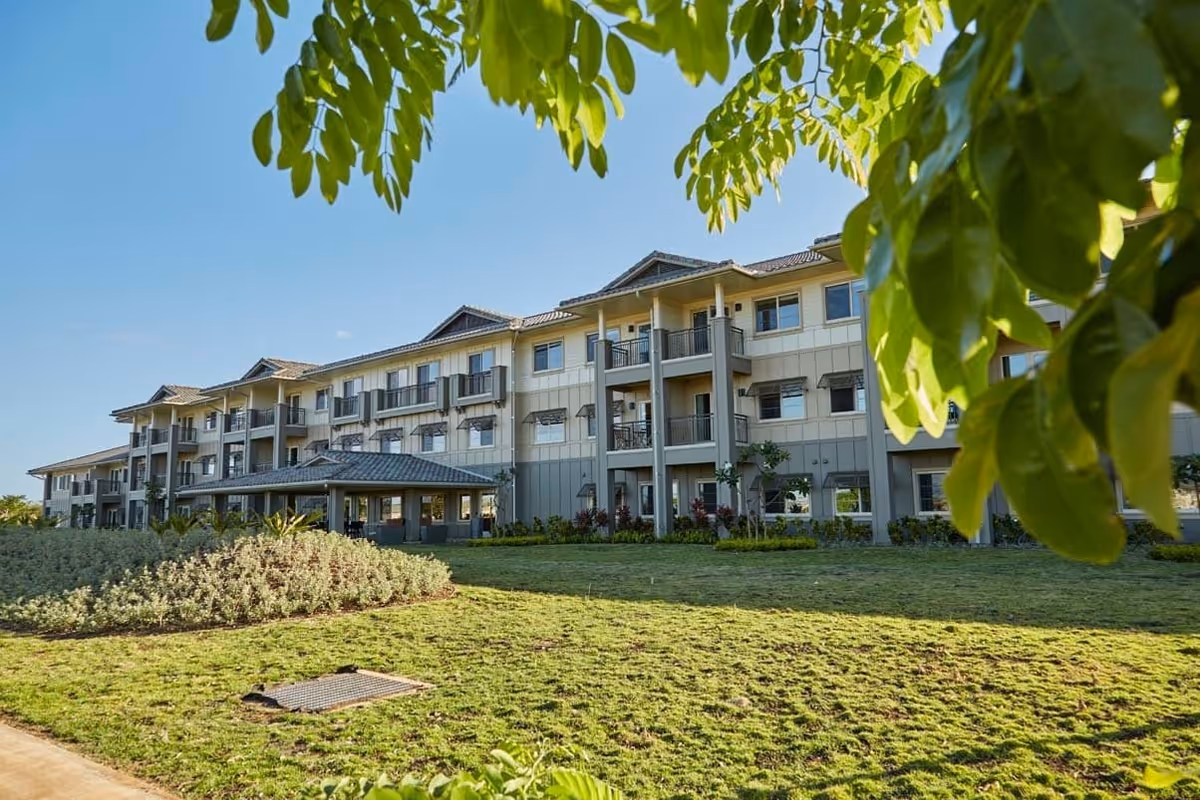 Exterior view of a three-story senior living facility building named Ilima at Leihano, surrounded by green grass and trees under a clear blue sky.