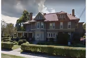 Large two-story brick house with a red tiled roof, multiple chimneys, and several windows. The house is surrounded by trimmed hedges and trees under a partly cloudy sky.
