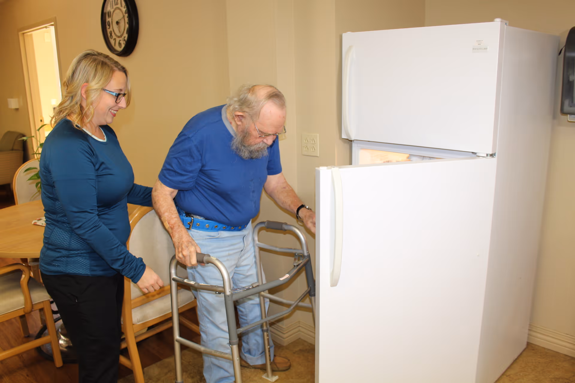 An elderly man using a walker is opening the door of a white refrigerator while a woman stands beside him, smiling and offering support. They are in a room with a wooden dining table and chairs, and a large wall clock is visible in the background.